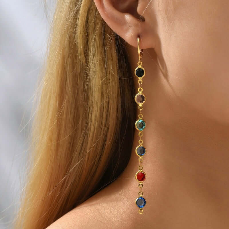 Gold earring with multicolored gemstones worn by a person with blonde hair.