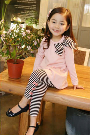 Adorable girl in pink striped bow tie shirt and leggings set, sitting at a wooden table with a plant.