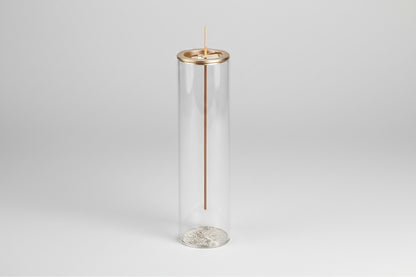 Clear cylindrical glass holder with a gold rim containing an incense stick on a white background
