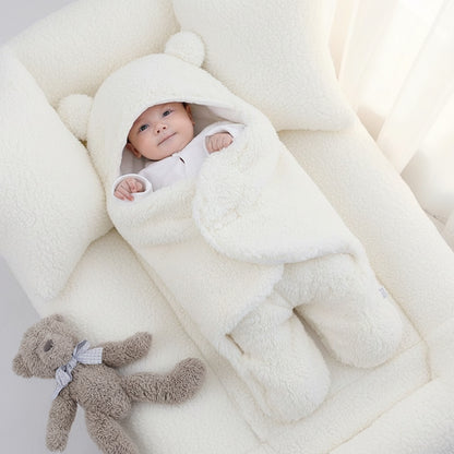 A baby wrapped in a white quilted sleeping bag with bear ears, lying next to a gray teddy bear toy.