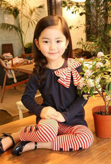 Adorable girl wearing a navy blue top with a striped bow and red striped leggings, sitting beside a potted plant.